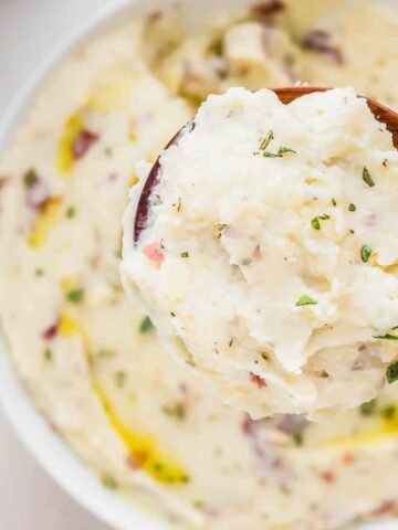 A wooden spoon holds a serving of creamy mashed potatoes with herbs, above a bowl filled with more mashed potatoes.