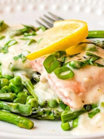 A plate of salmon topped with creamy white sauce, served with asparagus, peas, and garnished with lemon slices and chopped herbs. A fork rests on the plate.