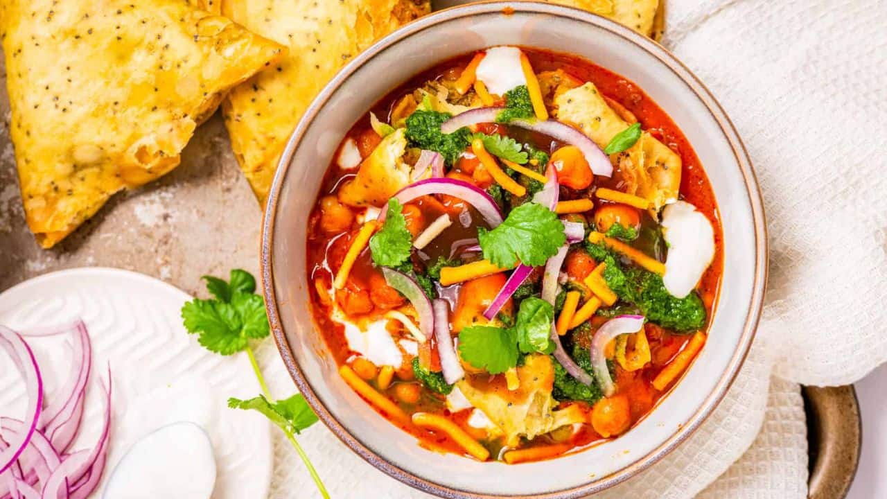 Straight view of samosa chaat served in a white bowl and is topped with cilantro, red onions, and sev. On the sides is a samosa and a cup of drink.
