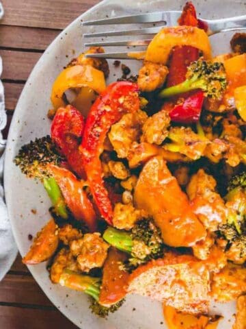 A plate of stir-fried chicken with broccoli, bell peppers, and carrots in sauce, served next to a baking tray on a wooden table.