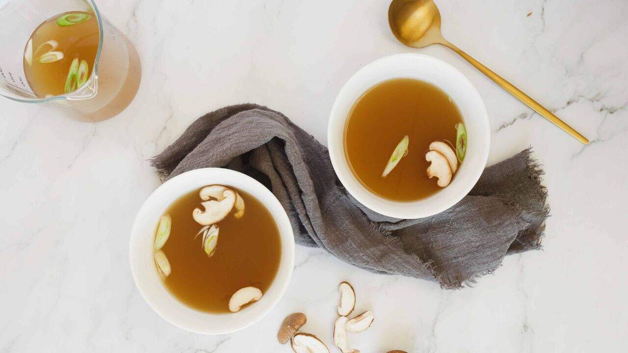 Two bowls of clear broth with sliced mushrooms and green onions on a white surface, next to a gray napkin, gold spoon, and a glass jug of broth.