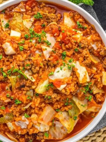 A bowl of cabbage roll casserole with ground meat, tomatoes, cabbage, and rice, garnished with herbs, sits on a checkered cloth next to an onion and woven placemat.