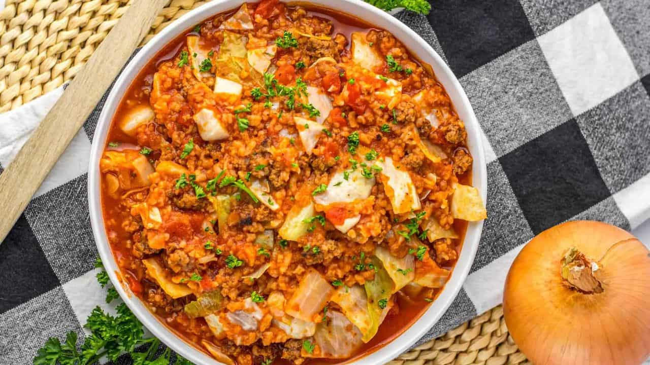 A bowl of cabbage roll casserole with ground meat, tomatoes, cabbage, and rice, garnished with herbs, sits on a checkered cloth next to an onion and woven placemat.