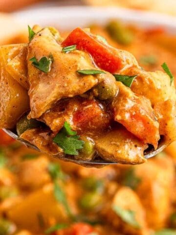 A close-up of a spoonful of vegetable curry with potatoes, peas, carrots, and sauce, garnished with chopped parsley.