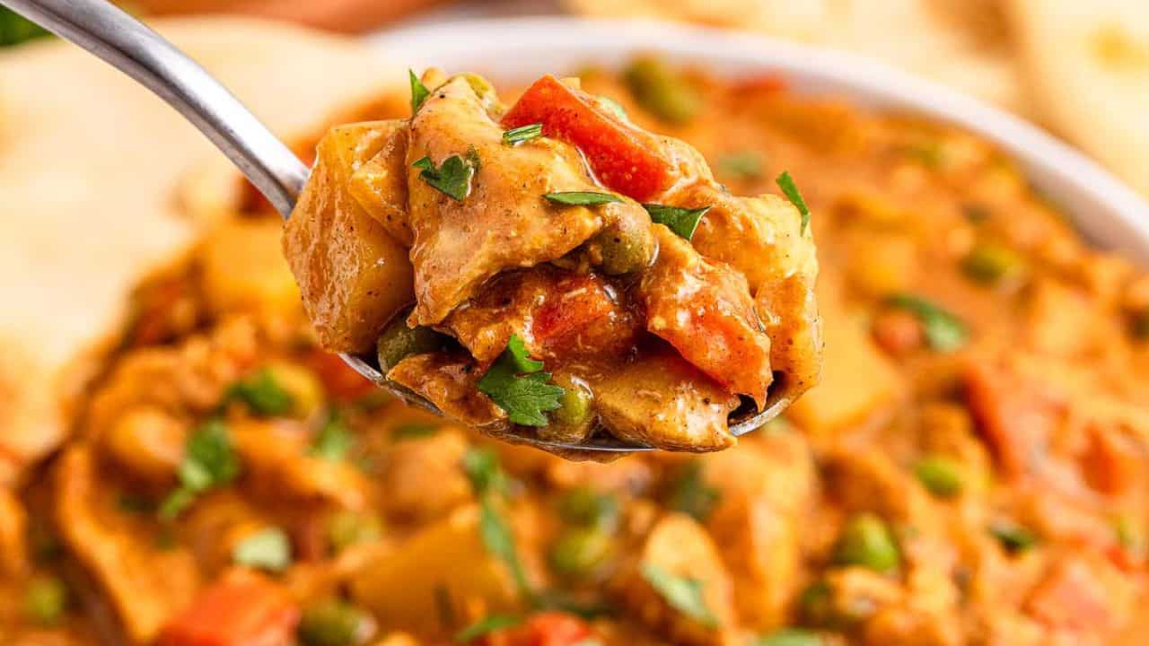 A close-up of a spoonful of vegetable curry with potatoes, peas, carrots, and sauce, garnished with chopped parsley.