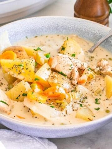 A bowl of creamy chicken stew with potatoes, carrots, and herbs, served with a spoon. Bread and parsley are visible in the background.