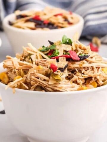 A white bowl filled with shredded chicken, corn, tortilla strips, and herbs sits on a table with other bowls containing similar ingredients.