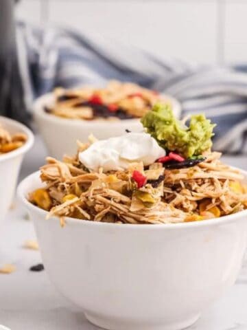 A bowl filled with shredded chicken topped with sour cream, guacamole, and tortilla strips, surrounded by ingredients like cheese, limes, and another bowl of chicken.