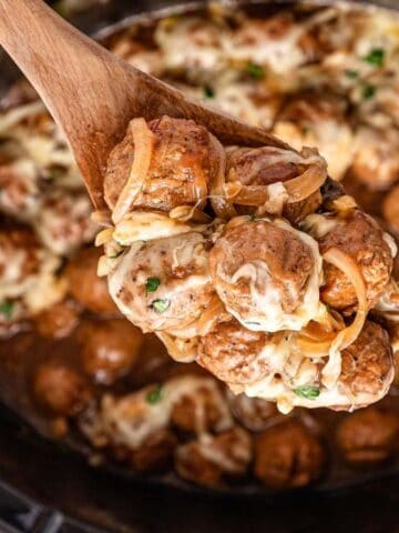 A wooden spoon holds cheesy meatballs and onions over a slow cooker filled with more meatballs in sauce.