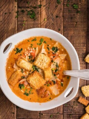 A bowl of creamy soup topped with croutons and herbs sits on a wooden table, accompanied by bread slices and additional bowls of soup.