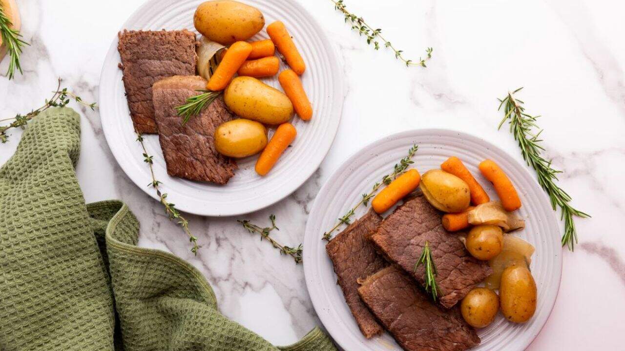 A plate of pot roast with sliced beef, baby potatoes, carrots, and onions, garnished with rosemary, sits on a table next to a green towel and a slow cooker.