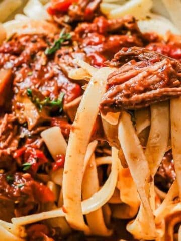 A bowl of fettuccine pasta topped with shredded meat and tomato-based sauce, with a fork twirling some pasta.