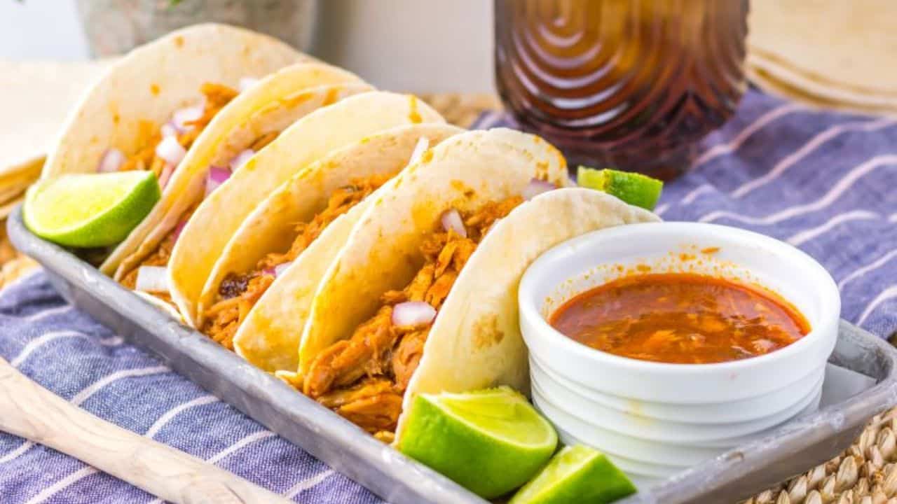 Tray of shredded pork tacos with diced onions and lime wedges, accompanied by a bowl of dipping sauce.