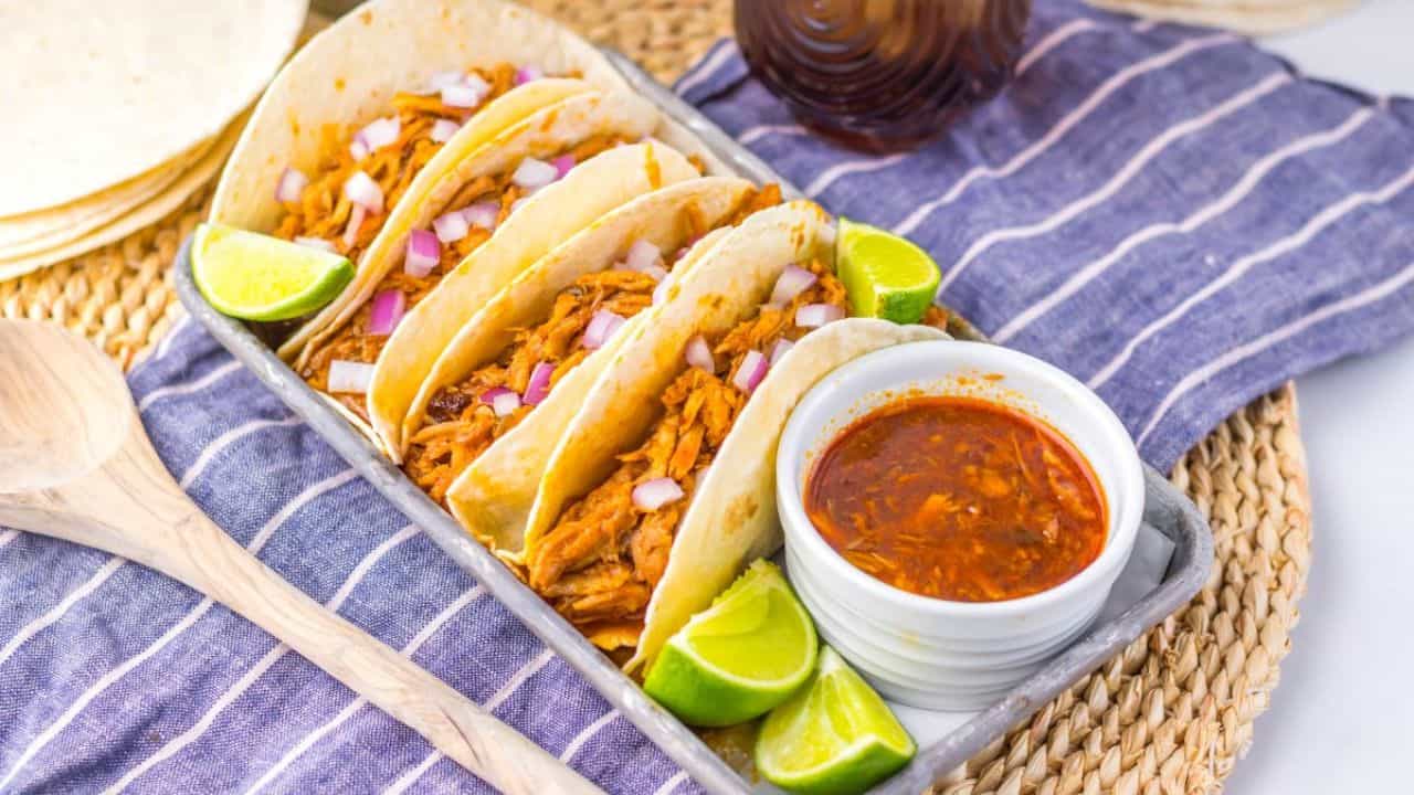Tray of shredded pork tacos with diced onions and lime wedges, accompanied by a bowl of dipping sauce.