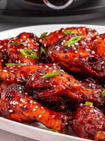 A white plate filled with glazed chicken wings garnished with sesame seeds and sliced green onions, with a small bowl of green onions on the side.