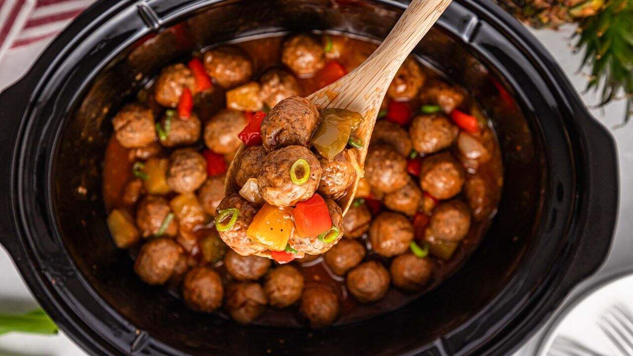A slow cooker filled with cooked meatballs, diced red and green peppers, and pineapple chunks, with a wooden spoon lifting a portion.