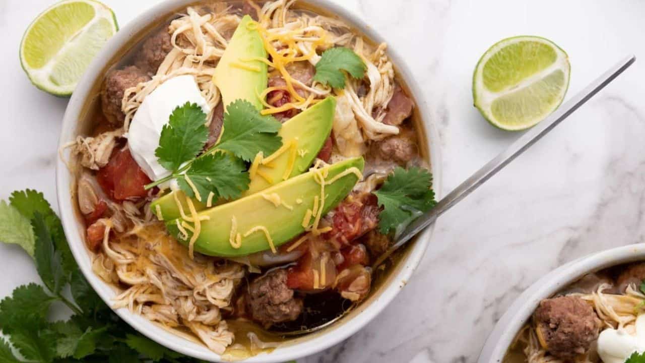 Two bowls of chili topped with avocado slices, sour cream, shredded cheese, and cilantro. Sliced lime and cheese bowl in the background on a marble surface.