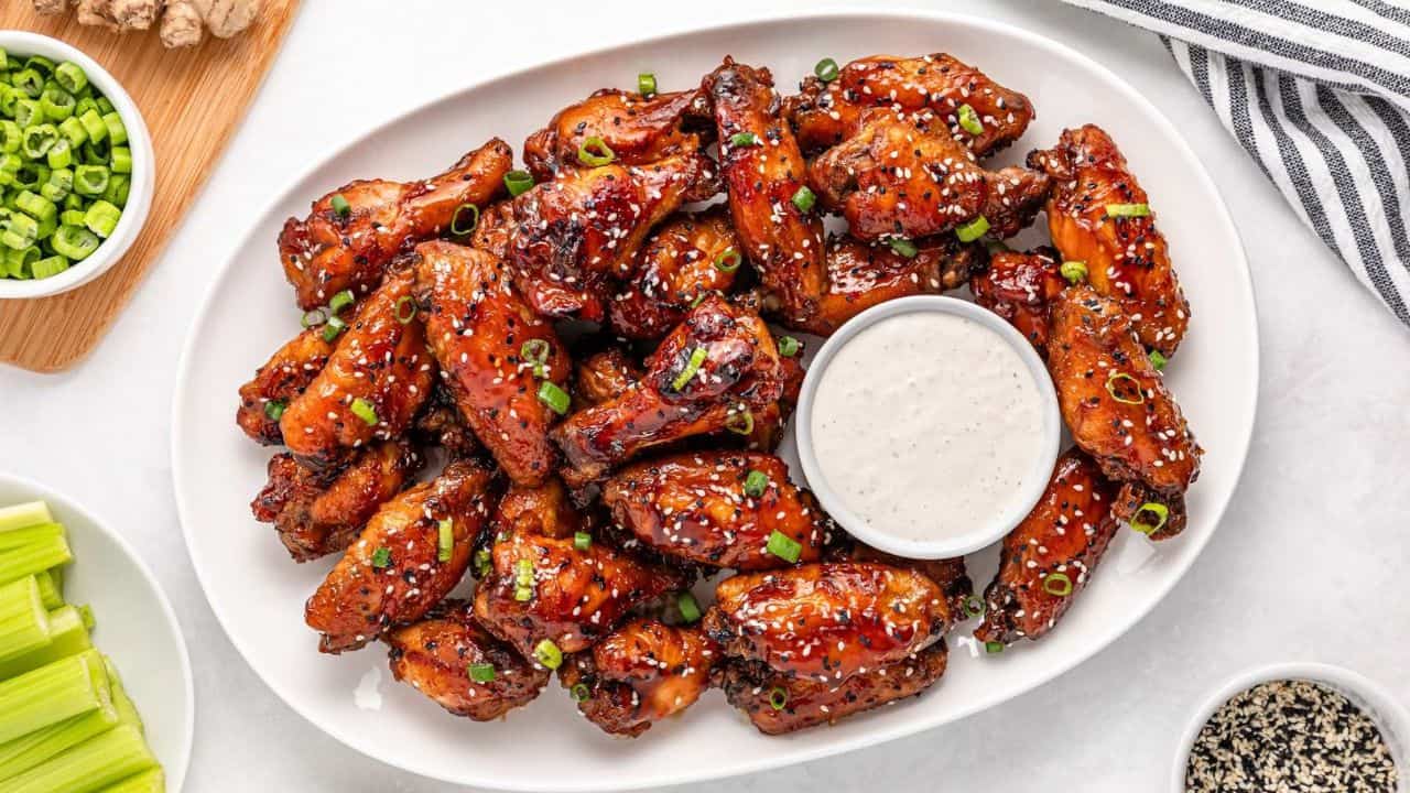 A platter of glazed chicken wings garnished with green onions and sesame seeds, served with a cup of ranch dip, celery sticks, and chopped green onions on the side.