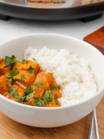A white bowl contains white rice and orange curry with herbs, placed on a wooden board. A spoon, naan bread, and a small bowl of chopped herbs are nearby.