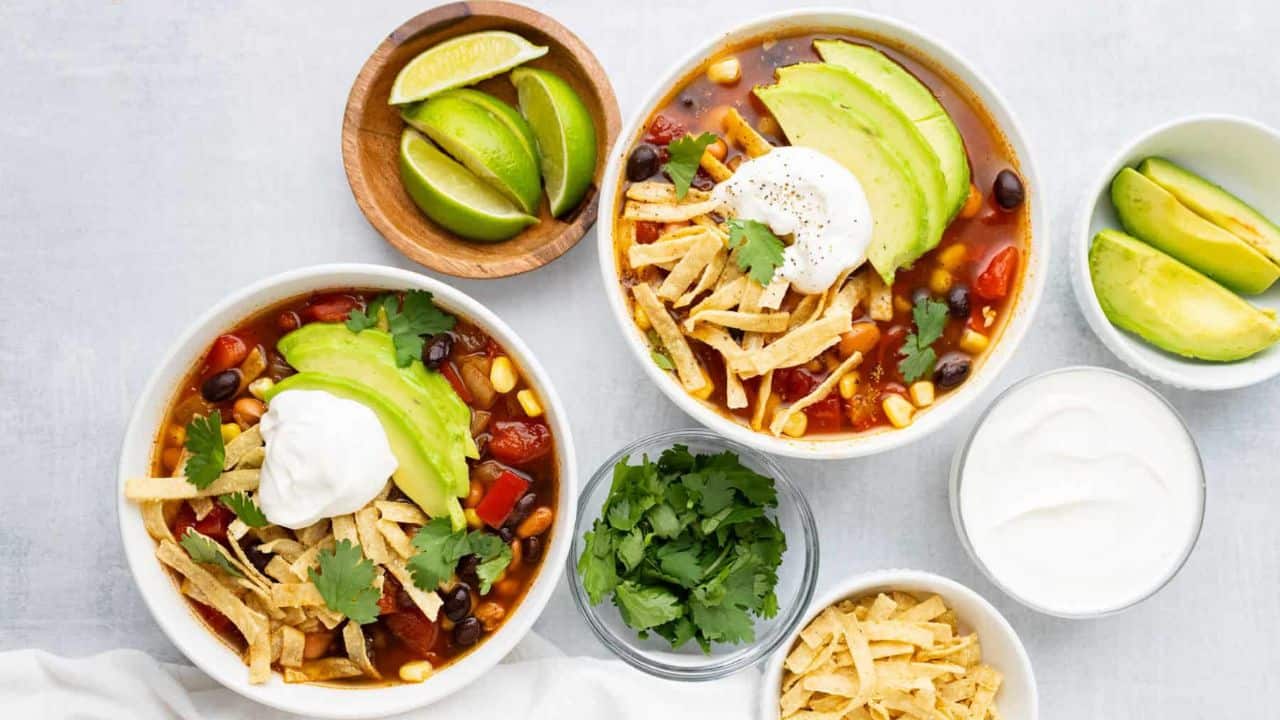 Two bowls of tortilla soup topped with avocado, sour cream, tortilla strips, and cilantro, surrounded by bowls of lime wedges, avocado slices, extra cilantro, sour cream, and tortilla strips.