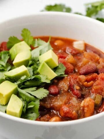 A white bowl of chili topped with diced avocado and fresh cilantro sits on a white surface, with a spoon and jalapeño pepper nearby.