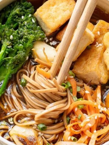 A bowl of noodle soup with tofu, broccolini, carrots, mushrooms, and sesame seeds, with chopsticks resting on top.