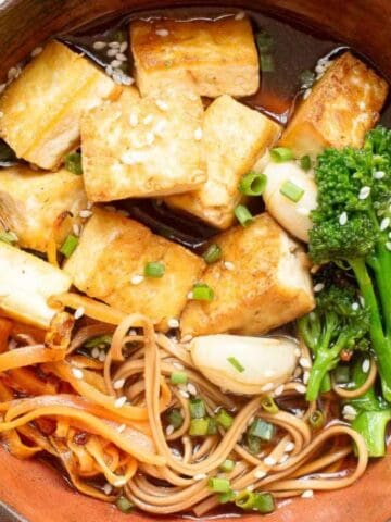 A bowl of tofu, soba noodles, broccoli, carrots, and green onions in broth, topped with sesame seeds.