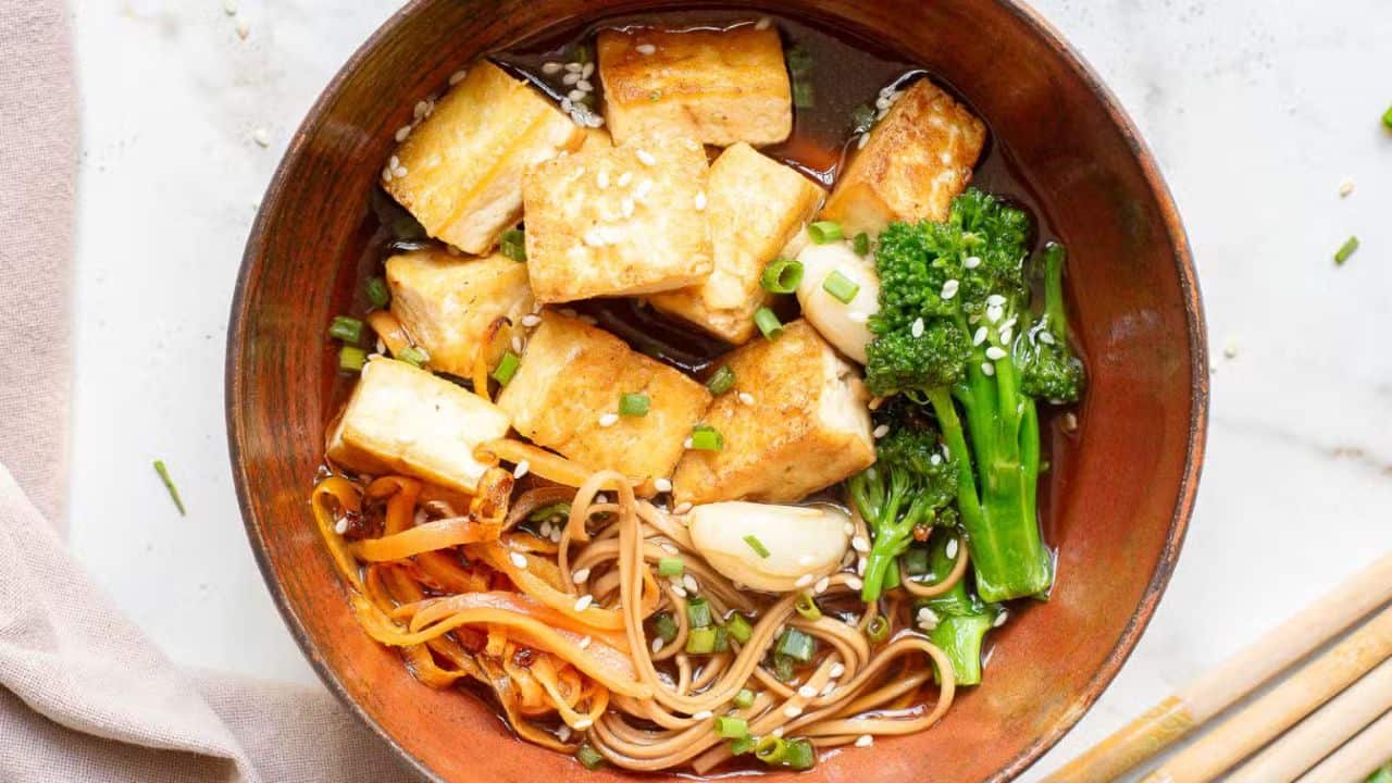 A bowl of soba noodles in broth, topped with fried tofu, broccolini, carrot spirals, and sliced mushrooms. Chopsticks rest on the bowl, and sesame seeds are sprinkled over the dish.