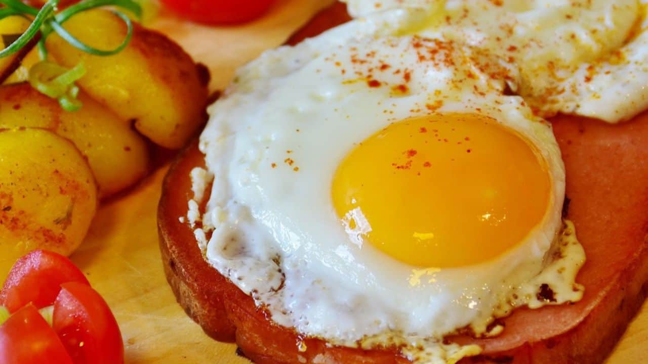A fried egg with a bright yellow yolk sits on a slice of ham, with roasted potatoes and sliced tomatoes on the side.