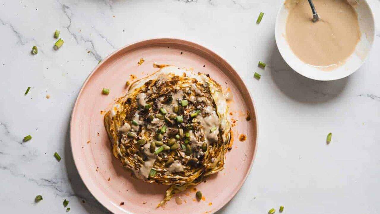 A roasted cabbage steak is served on a pink plate. It is topped with a creamy sauce, pumpkin seeds, and chopped green onions. The cabbage is browned and slightly charred at the edges.