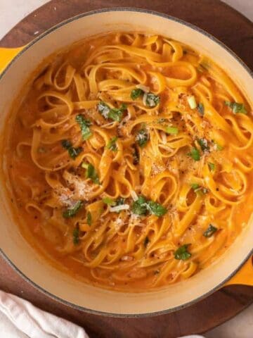 A pot of creamy tomato pasta garnished with herbs and grated cheese, placed on a wooden board next to a lid and a white kitchen towel.