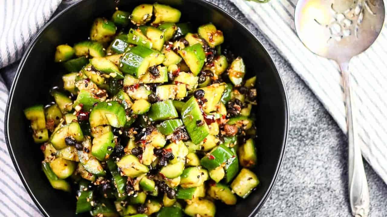 Overhead shot of spicy cucumber salad in a black bowl with a fancy serving spoon on the side.