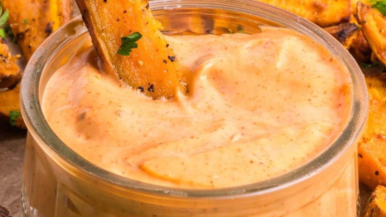 A glass bowl of creamy orange dipping sauce with a potato wedge partially dipped in it, surrounded by more potato wedges.