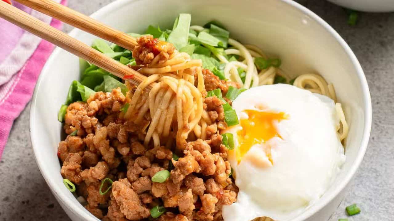 A bowl of noodles with minced meat, a poached egg, and chopped green onions. A hand uses chopsticks to lift the noodles. A purple cloth is partially visible on the side.