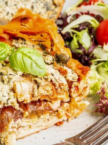 A slice of vegetable lasagna garnished with basil is served on a plate with a side salad of mixed greens and cherry tomatoes, next to a fork.