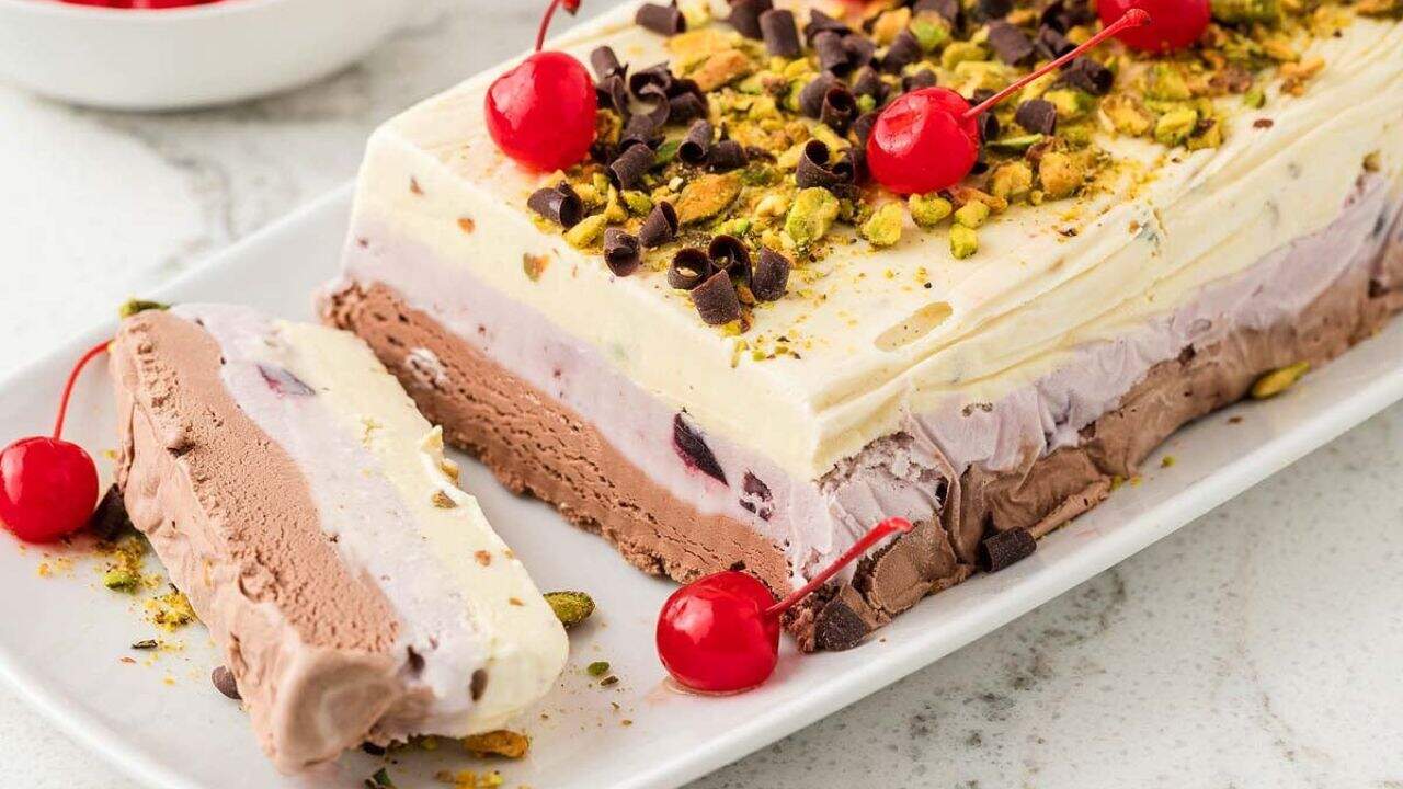 A sliced Neapolitan ice cream terrine topped with pistachios, chocolate curls, and cherries is served on a white platter.