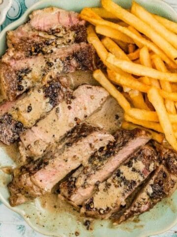A plate with sliced steak covered in peppercorn sauce and a serving of French fries, next to a fork and knife on a patterned surface.