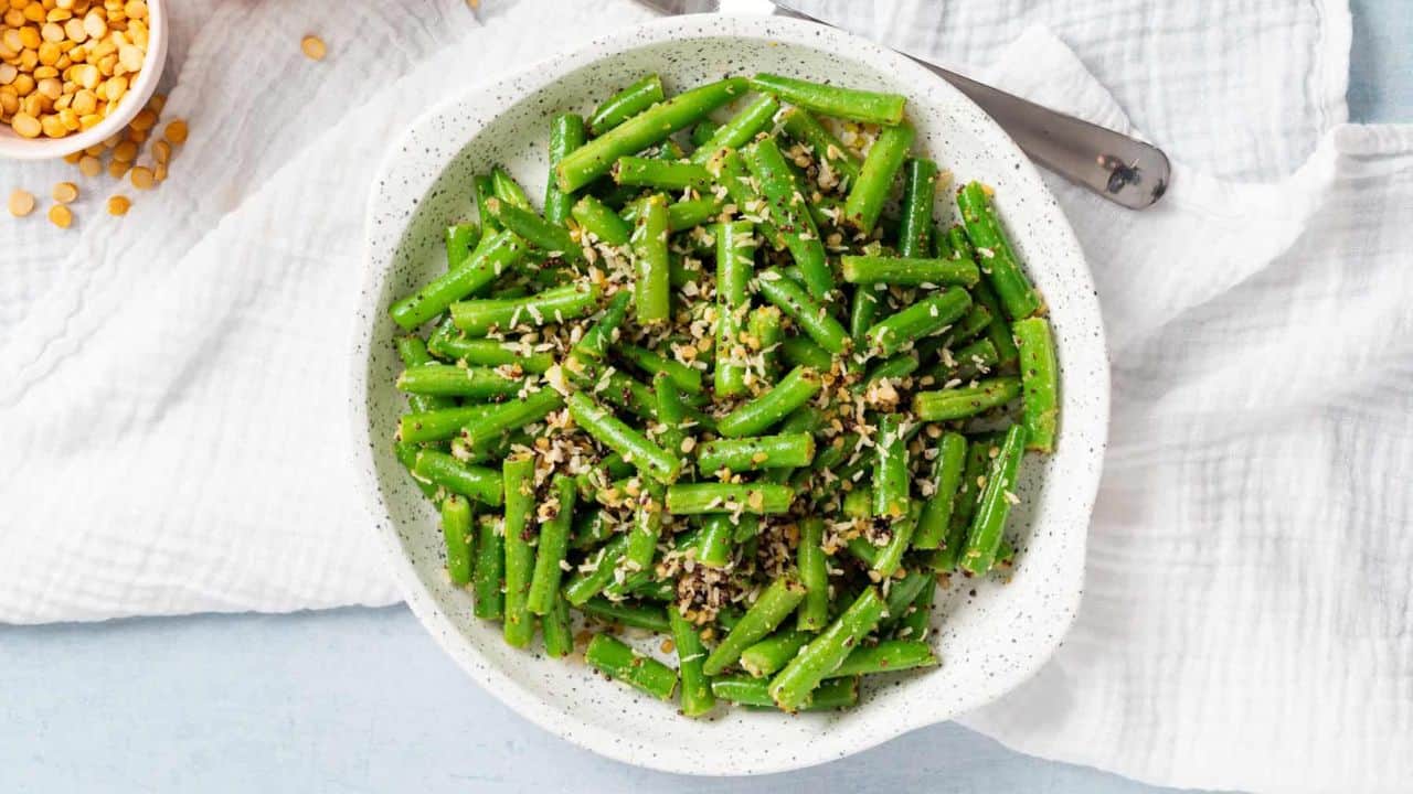 A top-down view of stir-fried green beans with coconut on a white plate, accompanied by a white napkin and fork.