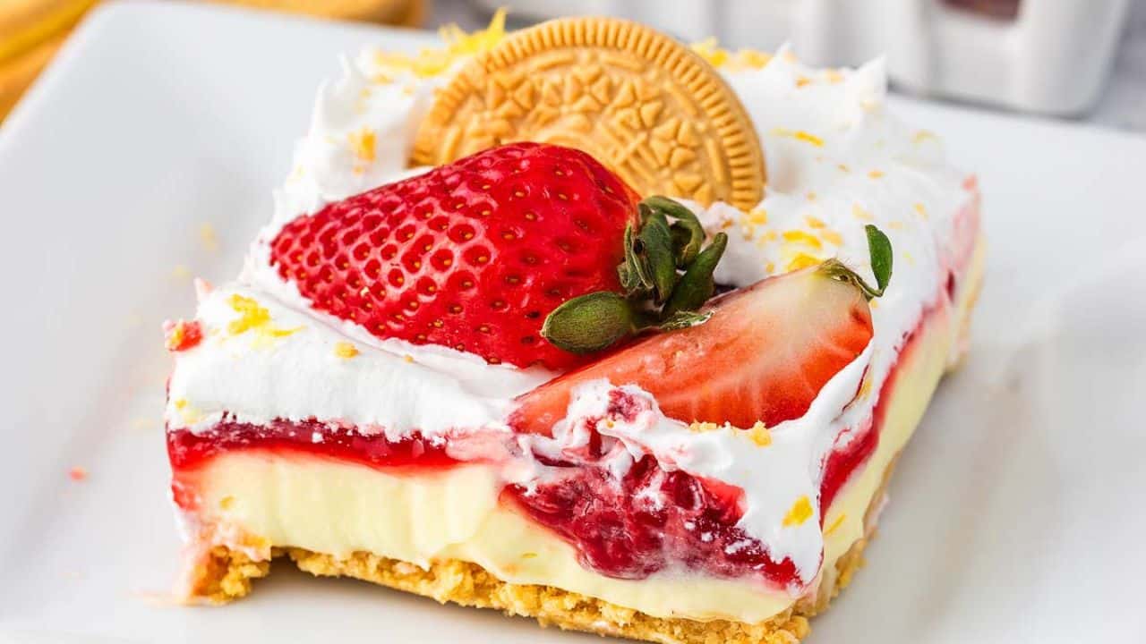 A dessert slice with layers of cream, strawberry topping, whipped cream, a whole strawberry, and a golden sandwich cookie on top, served on a white plate.
