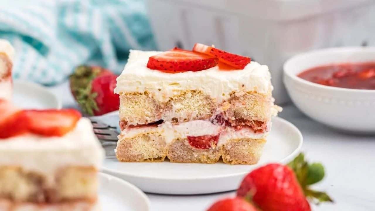 A slice of layered strawberry shortcake with cream and fresh strawberries on a white plate, with whole strawberries and a bowl of strawberry sauce nearby.