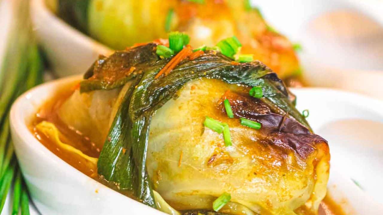 A close-up of a cooked cabbage roll topped with chopped chives, served in a white dish with sauce.