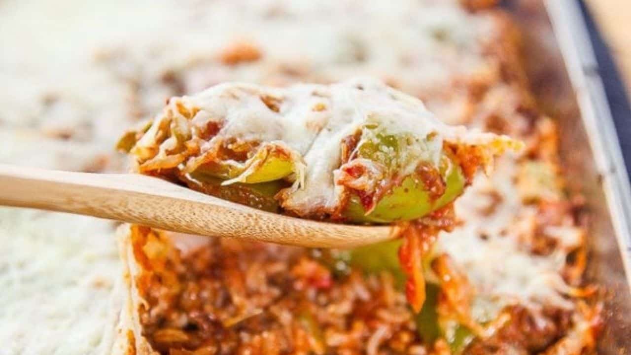 A wooden spoon lifts a serving of stuffed pepper casserole topped with melted cheese from a baking dish.