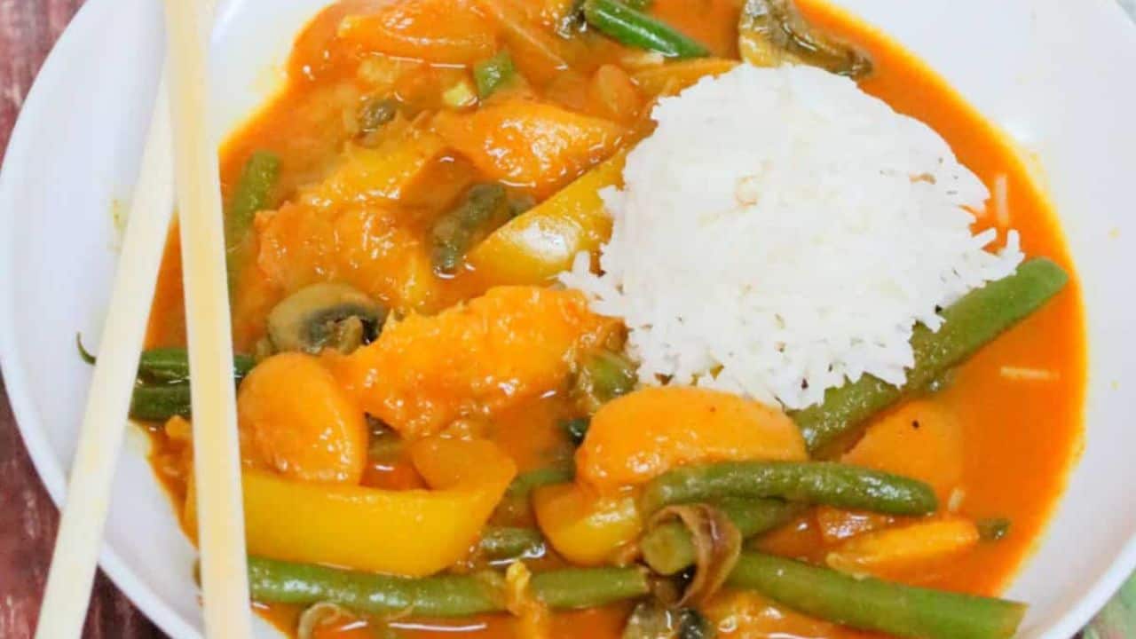A white bowl of curry with green beans, bell peppers, and mushrooms in an orange sauce, served with a portion of white rice and chopsticks on the side.