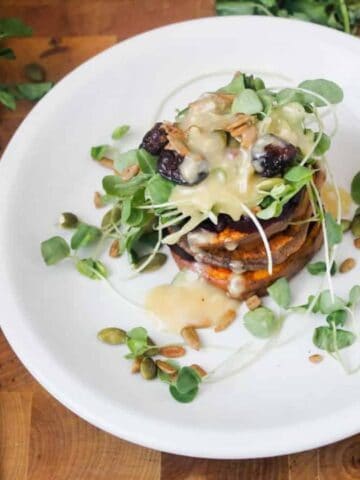 A neatly plated stack of roasted vegetables topped with microgreens and sauce on a white plate, with a knife and fork and fresh greens on a wooden surface.