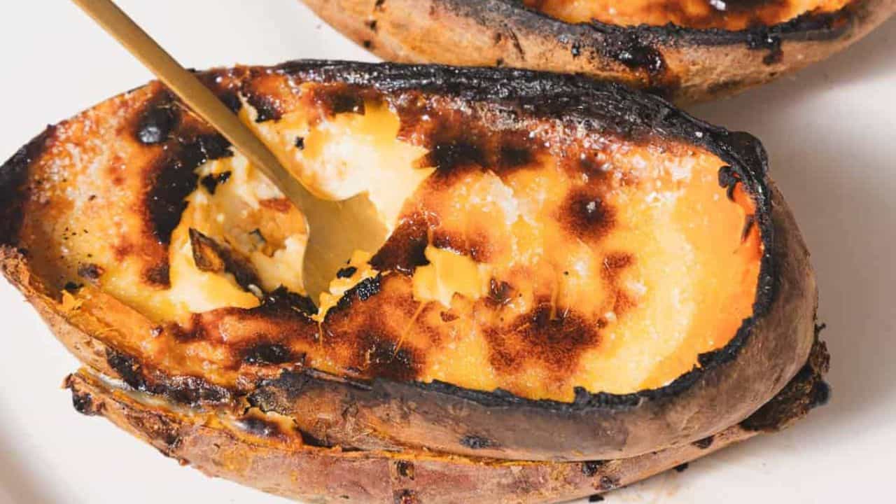 Two baked sweet potato halves on a white plate. The potatoes have a caramelized, slightly burnt surface, and one half is being scooped with a spoon.