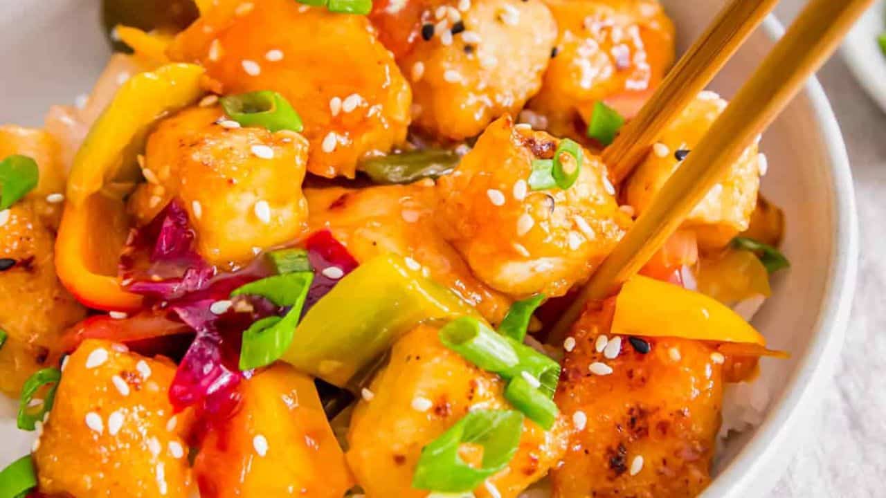 Sweet and sour tofu in a bowl with rice and chopsticks on the side.