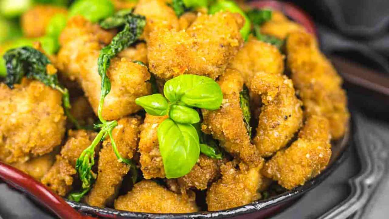 A bowl of crispy fried chicken with fresh basil leaves, accompanied by a side of sliced cucumbers and a basil in the background.