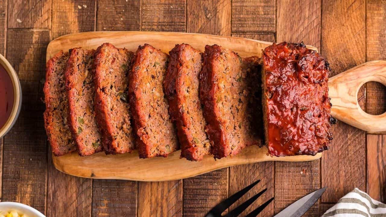 A sliced meatloaf with a glazed topping is arranged on a wooden cutting board on a wooden table.