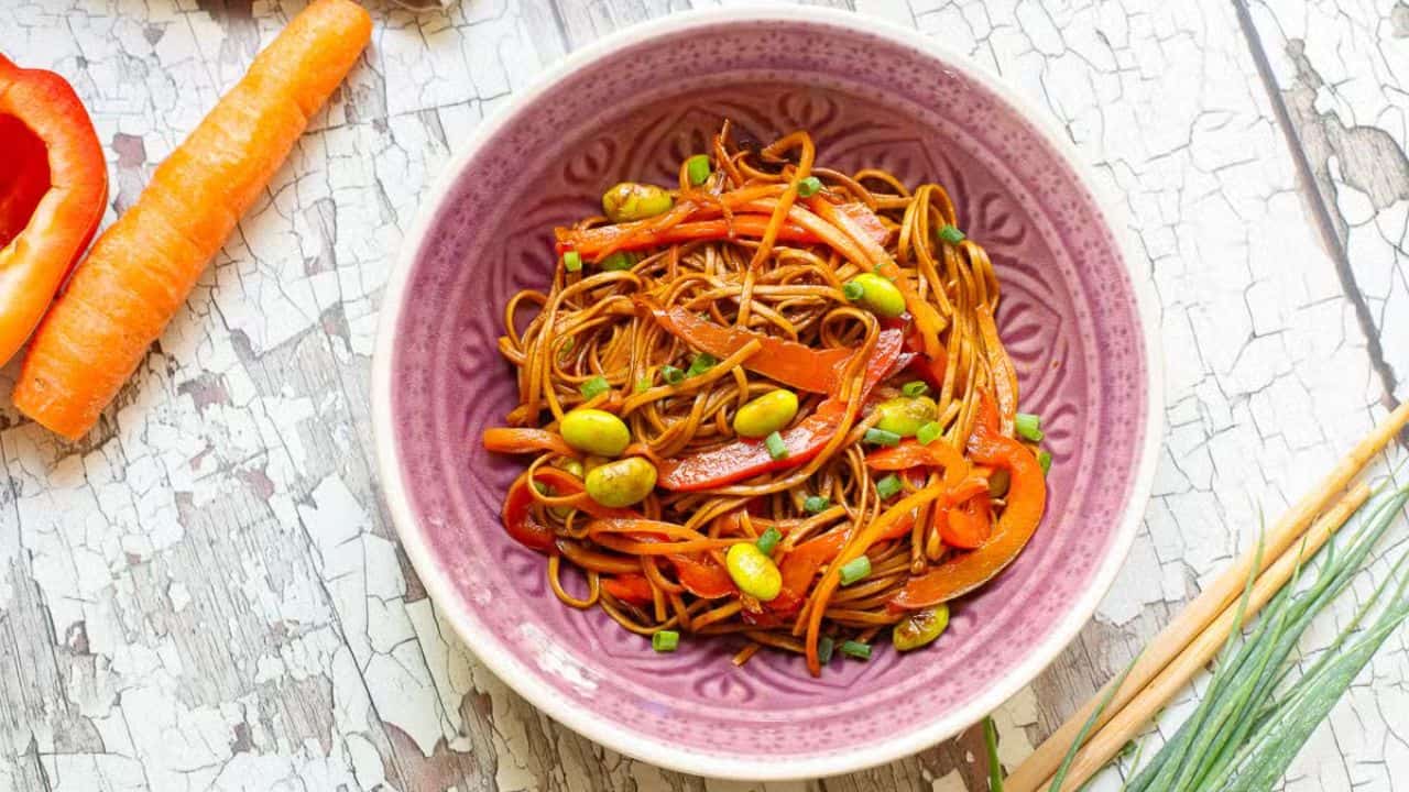 A purple bowl filled with noodles tossed with sliced red bell peppers, edamame, and green onions on a rustic white wooden surface. A few carrot slices and green onion leaves are partially visible on the side.