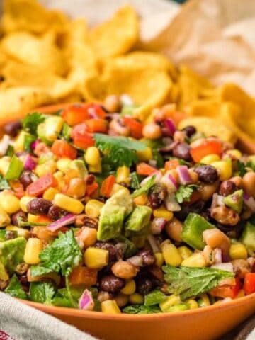 A bowl of colorful bean and corn salad with chopped vegetables is surrounded by a pile of corn chips on a tray lined with parchment paper.